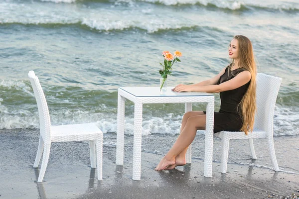A gyönyörű hölgy nyugszik a tenger kék háttér. Egy boldog lány egy fekete ruhát. Egy sor asztal és székek a strandon. — Stock Fotó