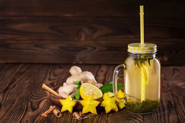 Γλυκό κίτρινο ποτό σε ένα μεγάλο γυάλινο βάζο με λεμόνι, κανέλα, μέντα, πιπερόριζα και καράμβολες σε φόντο ξύλινη. Χώρο αντίγραφο. — Φωτογραφία Αρχείου