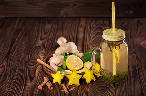 Bebida amarilla dulce en un gran frasco de vidrio de albañil con limón, canela, menta, jengibre y carambola sobre un fondo de madera. Copiar espacio . — Foto de Stock