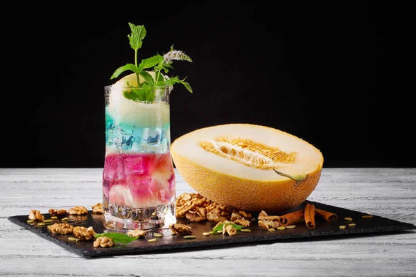 Eine frische saftige Melone und zerkleinerte Walnüsse neben einem bunten süßen Cocktail auf schwarzem Hintergrund. hellen Getränken. Kopierraum. — Stockfoto