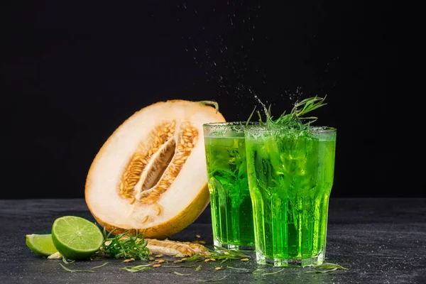 Two beverages and a melon on a black background. The green cocktails with lime and tarragon. Healthy, sweet and tasty drinks. Copy space.