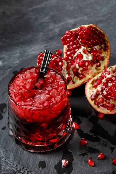 Un vaso grande de una bebida de granate sabrosa y fresca con hielo sobre un fondo negro. Granate fresco, maduro y natural roto sobre una mesa. Ingredientes ecológicos . — Foto de Stock