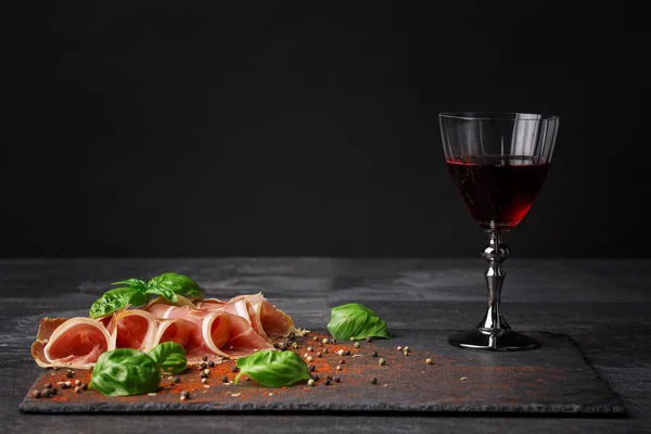 Una copa de vino y un montón de jamón cortado sobre un fondo negro. Prosciutto, albahaca verde fragante y granos de pimienta en el escritorio. Copiar espacio . — Foto de Stock