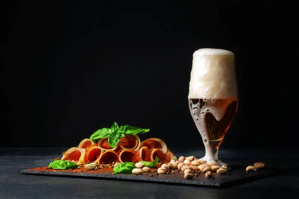 Ein großes Glas mit hellem Bier mit Schaum, köstlichem Schinken und Spinat auf schwarzem Hintergrund. Pistazien in der Nähe des Alkoholgetränks. salzige Snacks für Bier. — Stockfoto