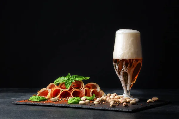 Um copo grande cheio de cerveja leve com espuma, delicioso prosciutto e espinafre no fundo preto. Pistache perto da bebida alcoólica. Petiscos salgados para cerveja . — Fotografia de Stock