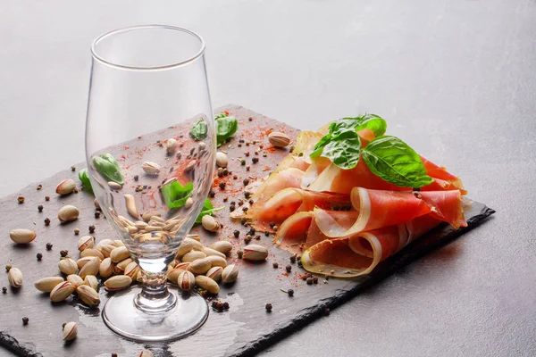 Ein leeres Glas, Basilikumblätter, Pistazien und rotgeschnittener Schinken auf hellgrauem Hintergrund. Leckere Snacks für ein alkoholisches Getränk. — Stockfoto