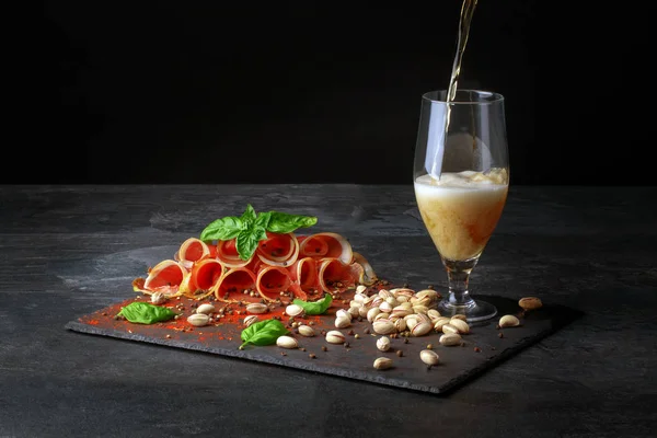 Ein großes Glas leichtes Bier und schmackhafter Prosciutto auf schwarzem Hintergrund. Pistazien in der Nähe des Alkoholgetränks. salzige Snacks für Bier. — Stockfoto