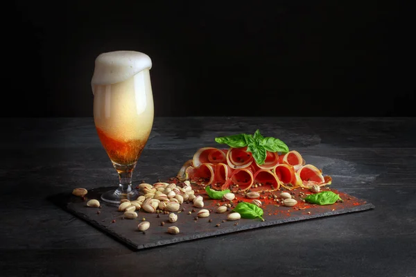 Ein großes Glas leichtes Bier und schmackhafter Prosciutto auf schwarzem Hintergrund. Pistazien in der Nähe des Alkoholgetränks. salzige Snacks für Bier. — Stockfoto