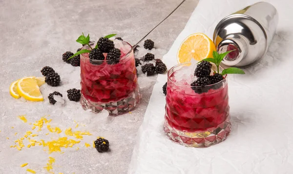 Blick von oben auf zwei große Gläser kalte Getränke mit Beeren und Eis auf grau-weißem Hintergrund. gesunde und biologische Brombeeren, Zitrone und Minze. — Stockfoto