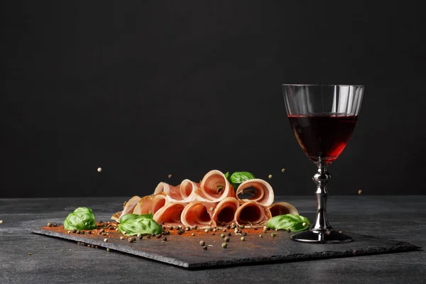 Ein Glas Rotwein und geschnittener Schinken mit Basilikum und Pfeffer auf schwarzem Hintergrund. lecker, gesund, köstlich und süß Wein. Kopierraum. — Stockfoto