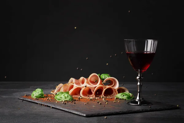 Ein Glas Rotwein und geschnittener Schinken mit Basilikum und Pfeffer auf schwarzem Hintergrund. lecker, gesund, köstlich und süß Wein. Kopierraum. — Stockfoto