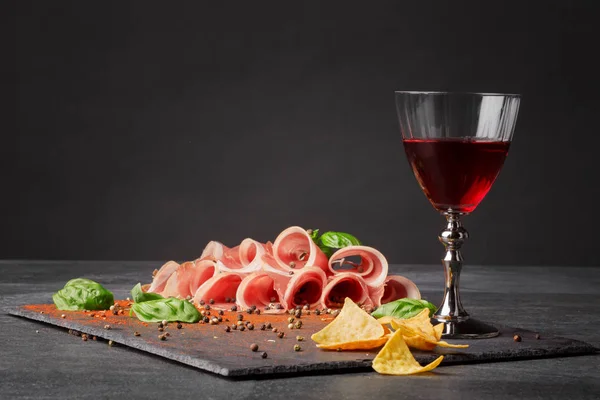 Zusammensetzung der Delikatesse. ein volles Weinglas, Nachos-Chips, roter Schinken und frischer Spinat auf schwarzem Hintergrund. Kopierraum. — Stockfoto