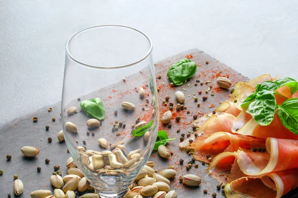 Um copo vazio, folhas de manjericão, pistache e presunto em fatias vermelhas em um fundo de bandeja leve. Petiscos saborosos para uma bebida alcoólica . — Fotografia de Stock