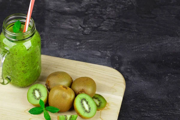 Ein Einmachglas gefüllt mit Kiwi-Getränk, Minzblätter auf einem hellen Holzbrett auf dunklem Steingrund. Kopierraum. — Stockfoto