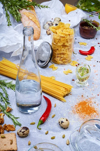 Close-up of a gray table with pasta, baguette, glass bottle, chili pepper, quail eggs and walnuts on a gray background. — Stock Photo, Image