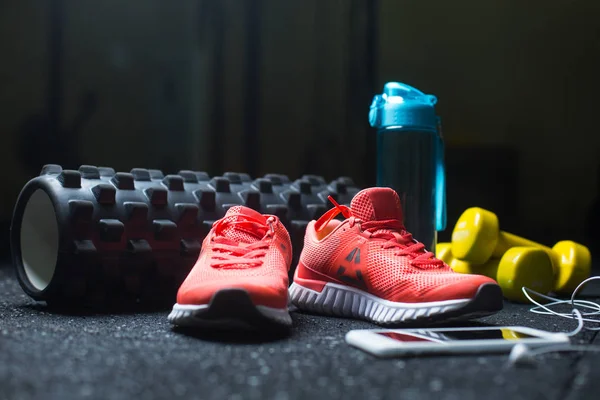 Crimson womens sneakers, yellow dumbbells, a bottle and a white mobile phone with headphones on a blurred background.