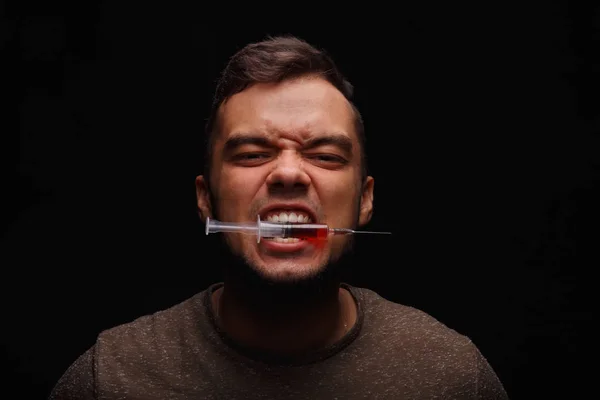 Um drogado com vícios de drogas prende entre os dentes uma seringa com uma droga injetável em um fundo preto escuro . — Fotografia de Stock