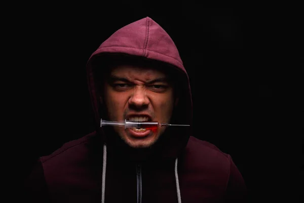 A junkie with drug addictions clamps between teeth a syringe with an injectable drug on a dark black background. — Stock Photo, Image