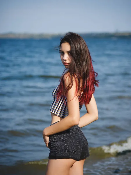 Confident young girl on a coast. Trendy, pretty girl model posing on a blurred water background. Teen fashion concept. — Stock Photo, Image