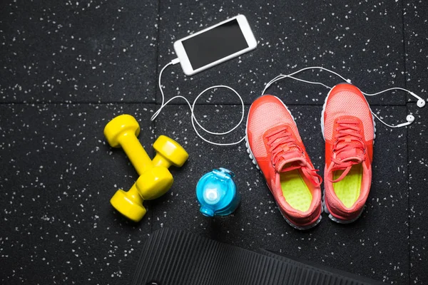 Top view of bright yellow dumbbells, a mat, bottle of water, sports shoes and phone on a black floor background. — Stock Photo, Image
