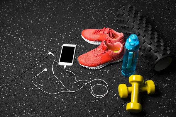 Comfortable training shoes, rubber mat, dumbbells, sports bottle and telephone on a black spotted background. Copy space.