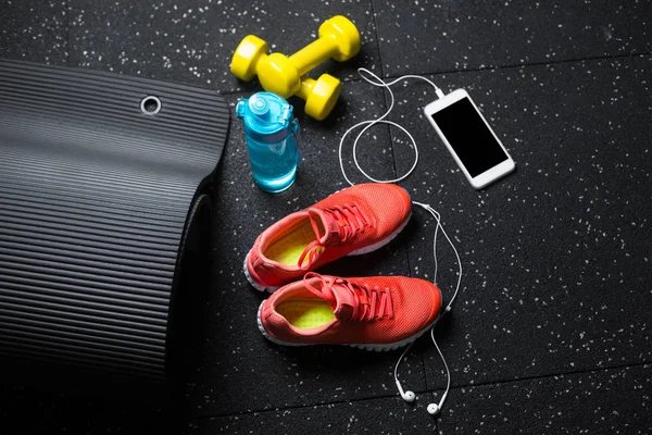 Top view of bright yellow dumbbells, a mat, bottle of water, sports shoes and phone on a black floor background.