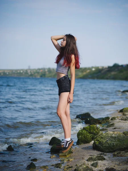 Zeker jonge meisje op een kust. Trendy, mooie meisje model poseren op een achtergrond wazig water. Teen fashion concept. — Stockfoto