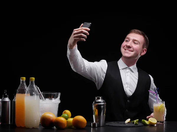 Привлекательный бармен на барной стойке делает селфи, шейкер, апельсины, лимон, лайм на черном фоне . — стоковое фото