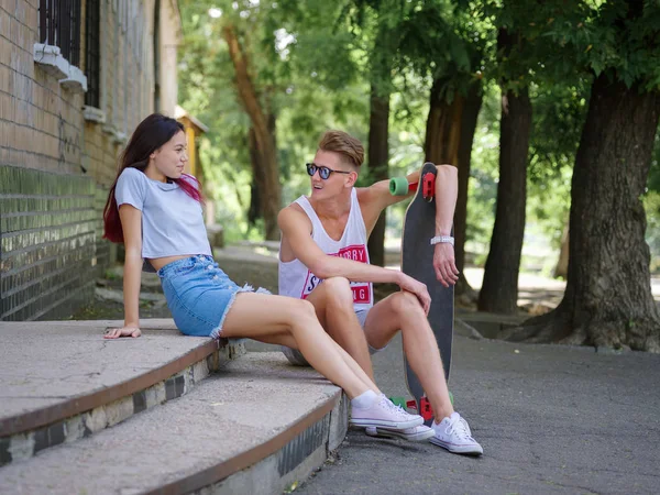 En vacker tjej och en kille med en longboard som sitter på sten trappor på en naturlig suddig bakgrund. — Stockfoto