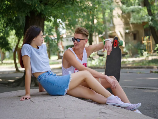 A beautiful girl and a fellow with a longboard sitting on stone stairs on a natural blurred background. — Stock Photo, Image