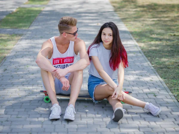 Teens dating in a park, a beautiful girl and a fellow sitting on a longboard on a natural blurred background. — Stock Photo, Image