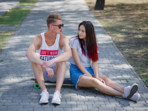 Adolescentes citas en un parque, una hermosa chica y un compañero sentado en un longboard sobre un fondo borroso natural . — Foto de Stock