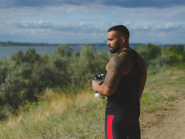 Egy izmos, jóképű testépítő egy tetoválás a váll ivóvíz a természetes háttér edzés után a. — Stock Fotó