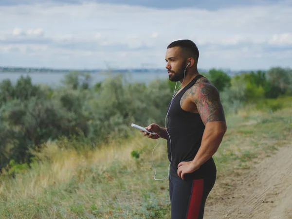 Egy izmos, jóképű bodybuilder a tetoválás a vállán hallgat zenét a természetes háttér edzés után a. — Stock Fotó