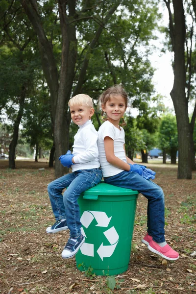 Niños espalda con espalda en pantalones vaqueros y pantalones cortos blancos, sentados en la gran papelera de reciclaje verde con etiqueta blanca en la parte delantera . — Foto de Stock