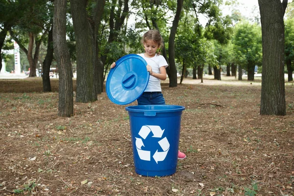 En liten söt flicka öppnar en mörk blå återvinning bin på en suddig naturliga bakgrund. Ekologi och barn. — Stockfoto