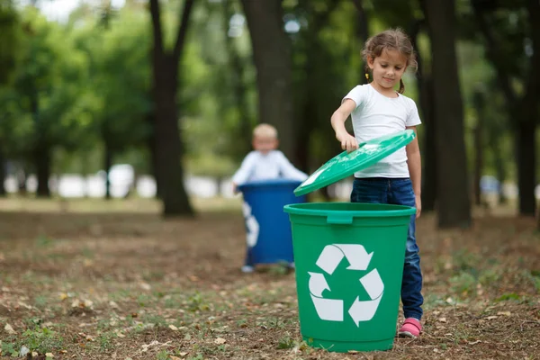 Una niña poniendo una tapa de cubo en una papelera de reciclaje verde sobre un fondo natural borroso. Ecología y niños . — Foto de Stock