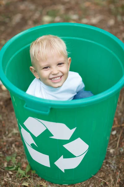 Um menino louro bonito sentado em uma lixeira verde em um fundo de chão de parque borrado. Conceito de poluição ecológica . — Fotografia de Stock