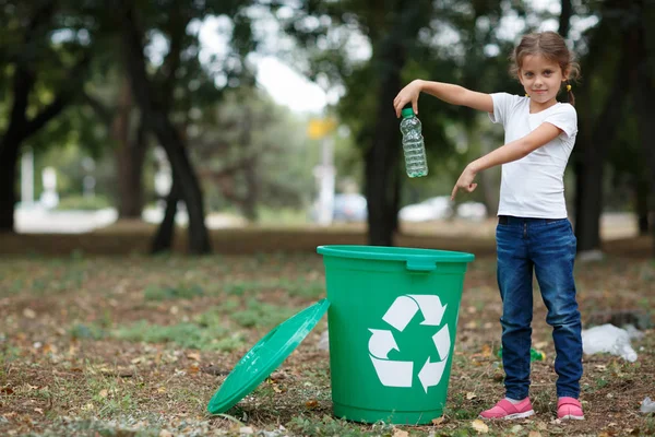 Uma criança colocando o lixo em uma lixeira verde em um fundo natural desfocado. Conceito de poluição ecológica . — Fotografia de Stock