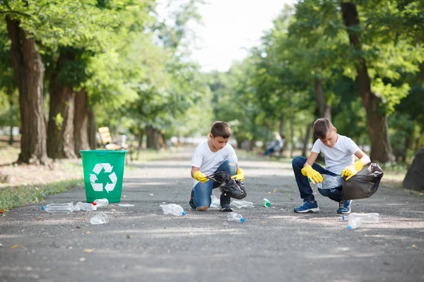 Két kis környezetvédők ül és gyűjtése műanyag szemetet, a homályos park háttér. Ökológiai védelmi koncepció. — Stock Fotó