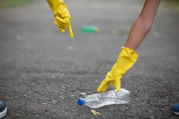 Childs händer i gul latex handskar plocka upp en plastflaska på en asfalt bakgrund. Ekologi skydd koncept. — Stockfoto