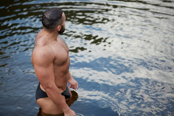 Un hombre musculoso sexy en bañadores mirando a la distancia en la orilla del mar. Un nadador sobre un fondo de agua . — Foto de Stock