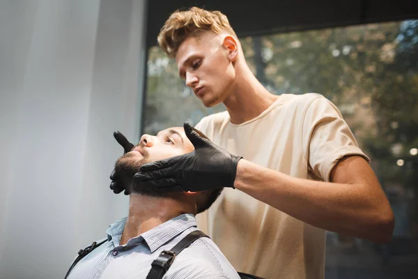 Krásný muž, sedící v křesle holiče a oholil holiče na pozadí salon krásy. Holičství koncepce. — Stock fotografie