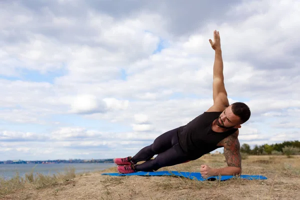 Un hombre deportivo con un cuerpo musculoso haciendo ejercicios sobre una alfombra en un fondo natural. Concepto de deportes y yoga . — Foto de Stock