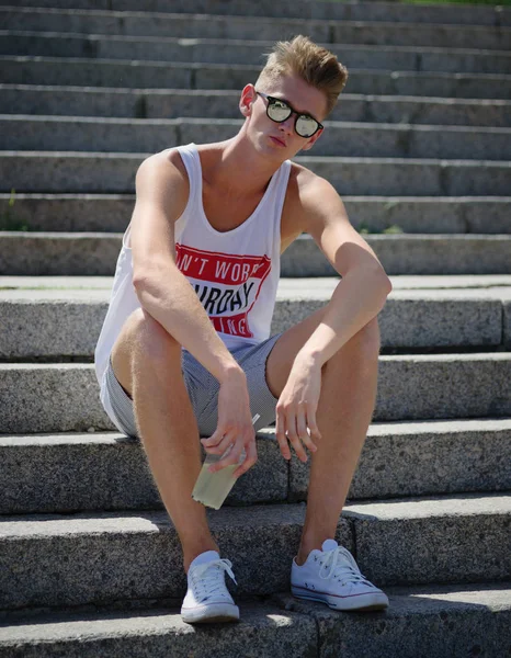 Ein Jugendlicher in weißem T-Shirt und kurzer Hose mit Limonade auf Steintreppen-Hintergrund. Freizeit-, Dynamik- und Modekonzept. — Stockfoto