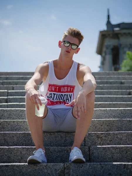 Um jovem em uma camiseta branca e shorts com uma limonada em um fundo natural. Recreação, juventude e conceito de moda . — Fotografia de Stock