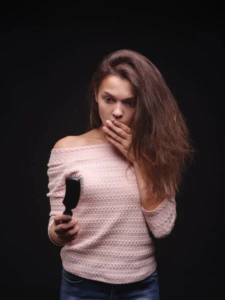 Ung kvinna med hår hälsa problemen ser orolig på kammen på svart bakgrund. Hair care koncept. Kopiera utrymme. — Stockfoto