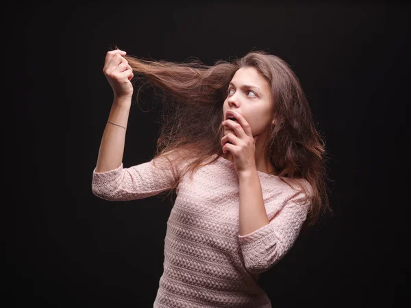 Una mujer bonita y preocupada tocando el pelo sobre un fondo negro. Mujer joven decepcionada con su cabello. Concepto de cabello frágil . — Foto de Stock