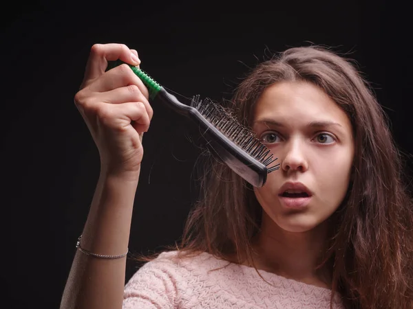 Menina lutando com cabelos emaranhados no fundo preto. Mulher de close-up olhando para um pente com cabelo. Conceito calvo . — Fotografia de Stock