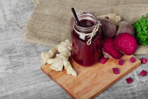 A mason jar of beetroot juice with black straw on a wooden background. A ginger, beets, and parsley on a cutting board.